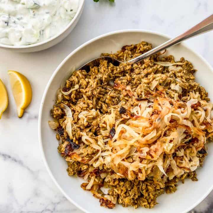 A large bowl of mujaddara next to a bowl of yogurt sauce and sliced lemons.