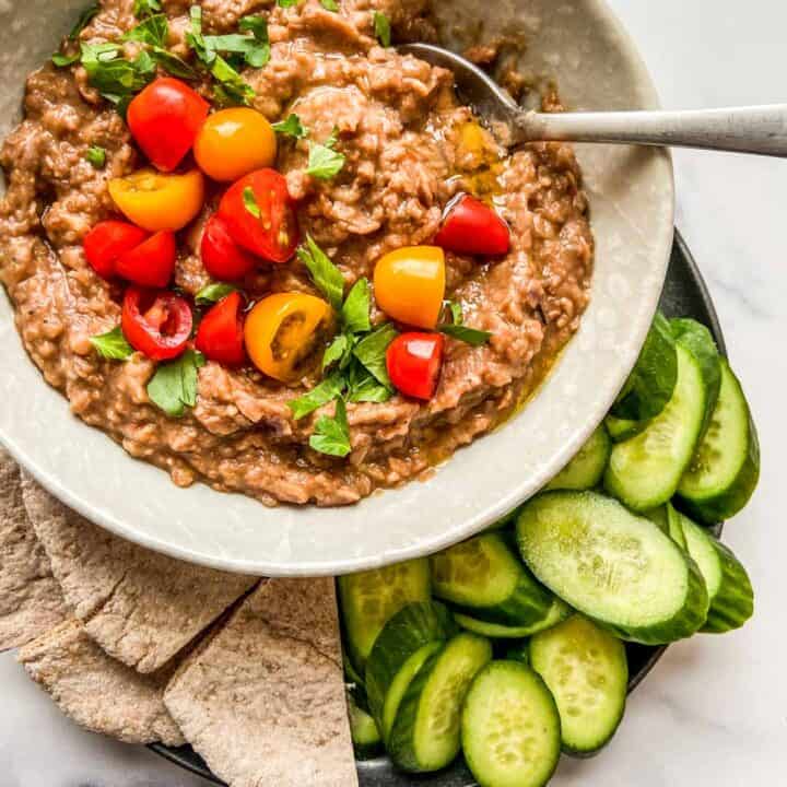 A bowl of ful medames topped with tomatoes and parsley, next to cut pita bread and sliced cucumbers.