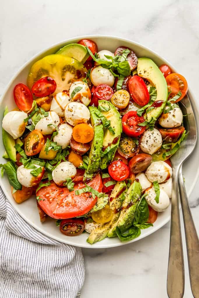 Avocado caprese salad in a large white bowl with serving spoons.