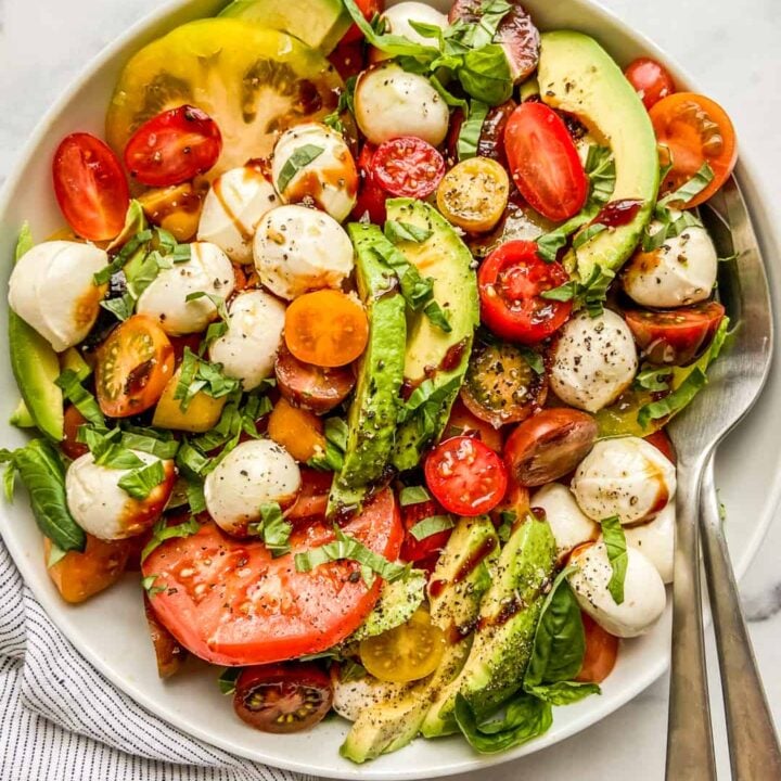 Avocado caprese salad in a large white bowl with serving spoons.