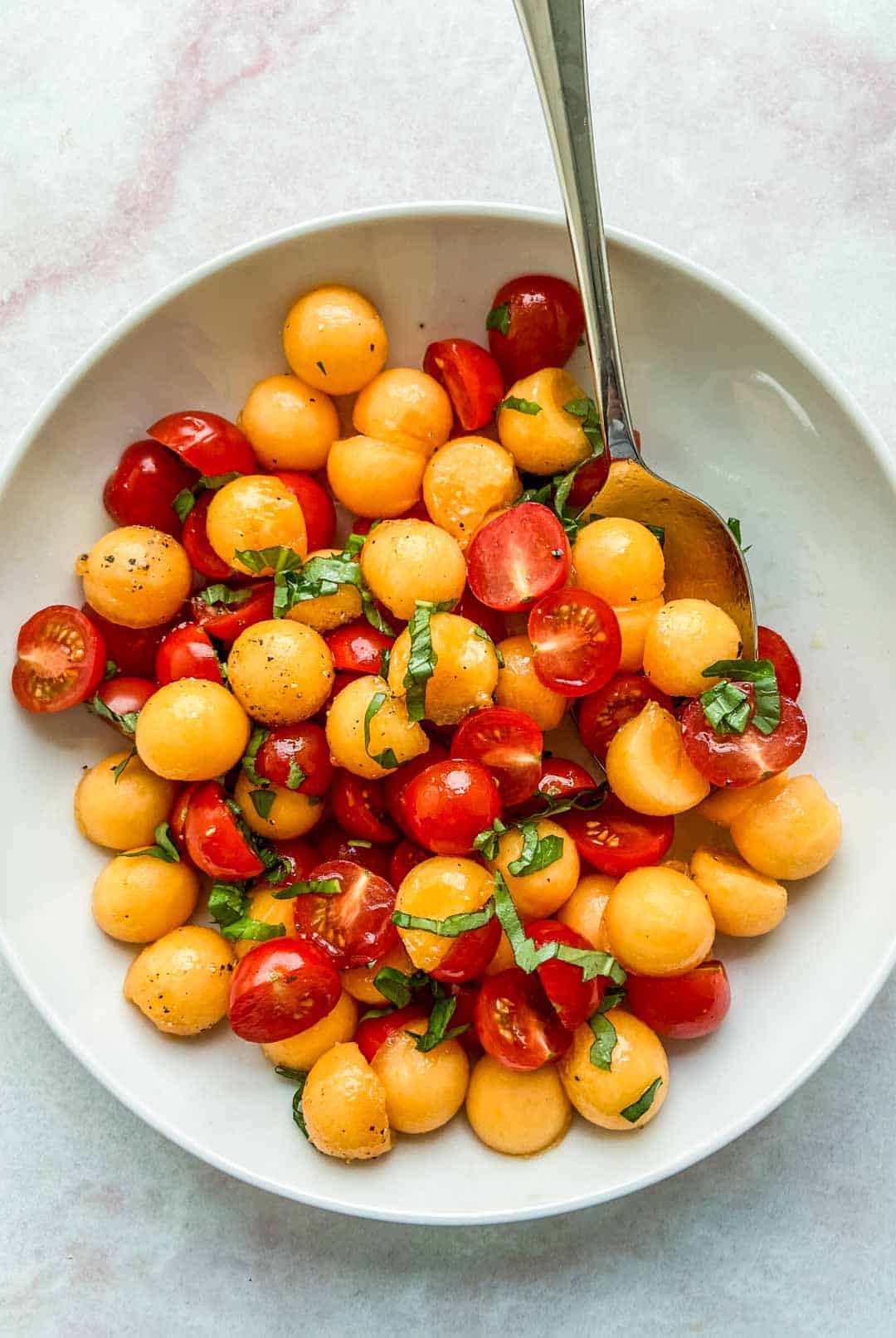 Tomato and cantaloupe salad in a white bowl with a serving spoon.