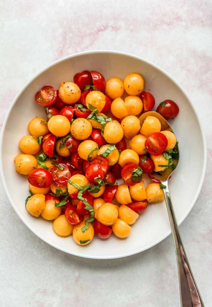 cherry tomato and cantaloupe salad in a white serving bowl with a spoon