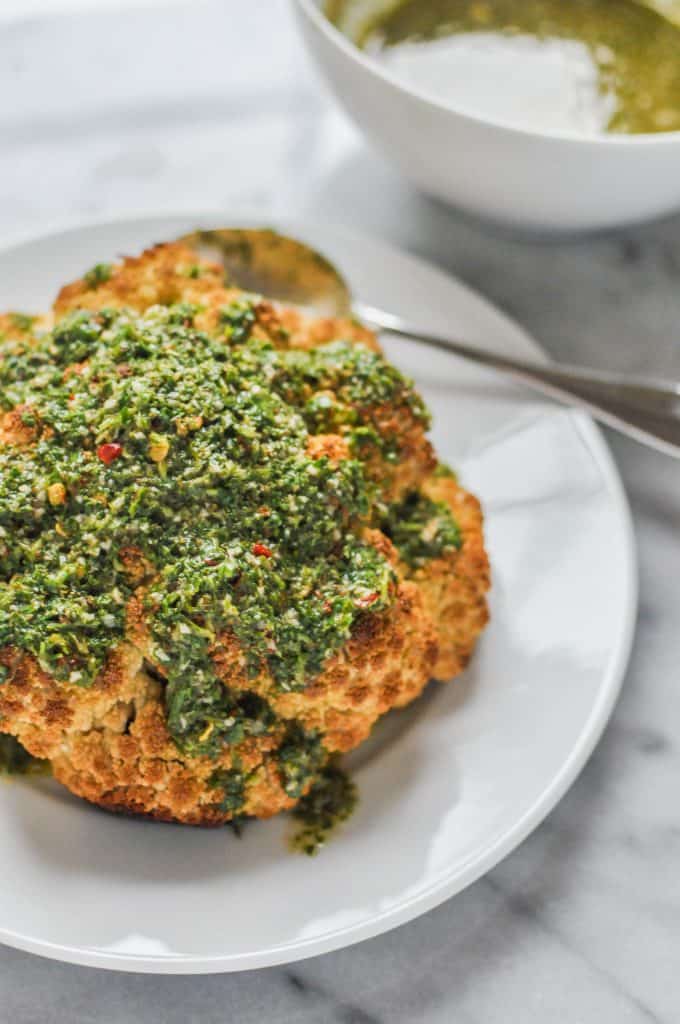 Whole roasted cauliflower with chimichurri sauce on a white plate.