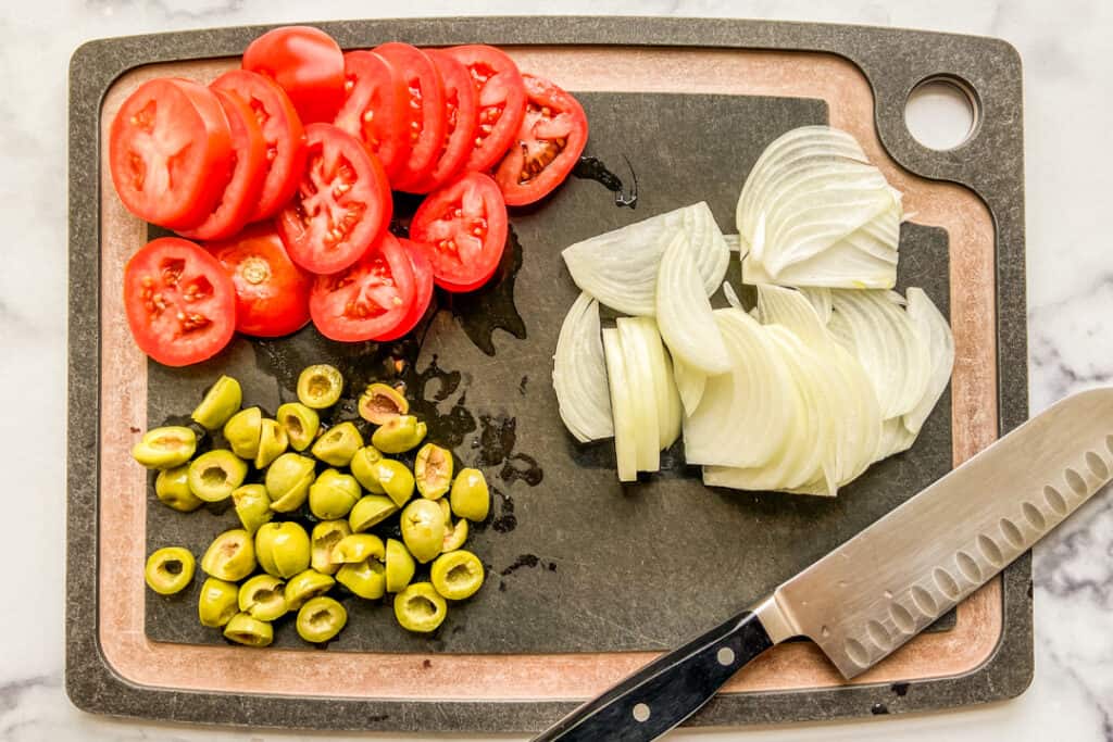 Cut tomatoes, olives, and onions on a cutting board.