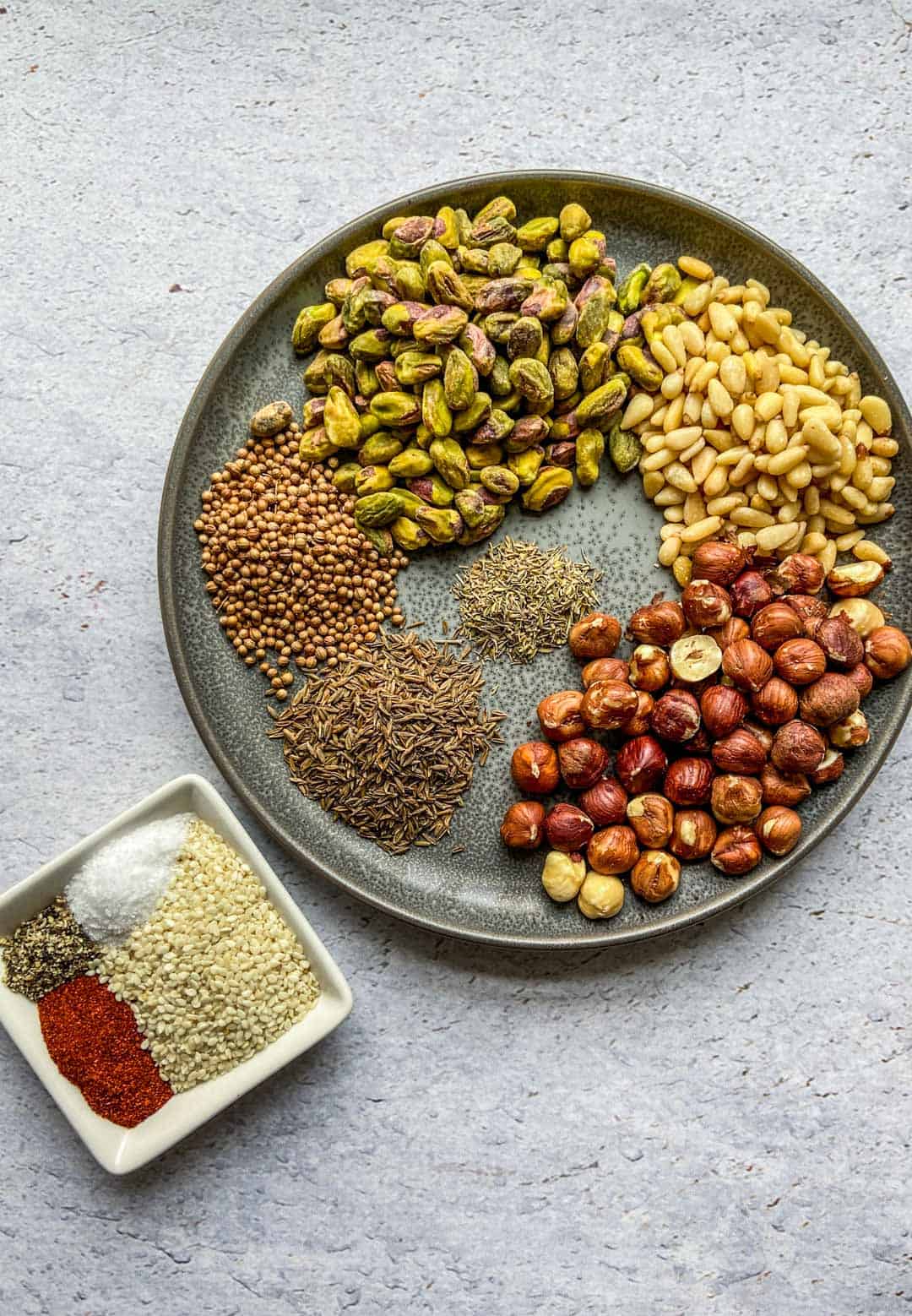 Pine nuts, hazelnuts, cumin seeds, coriander seeds, thyme, and pistachios on a plate, with sesame seeds, paprika, salt, and pepper in a little ramekin.