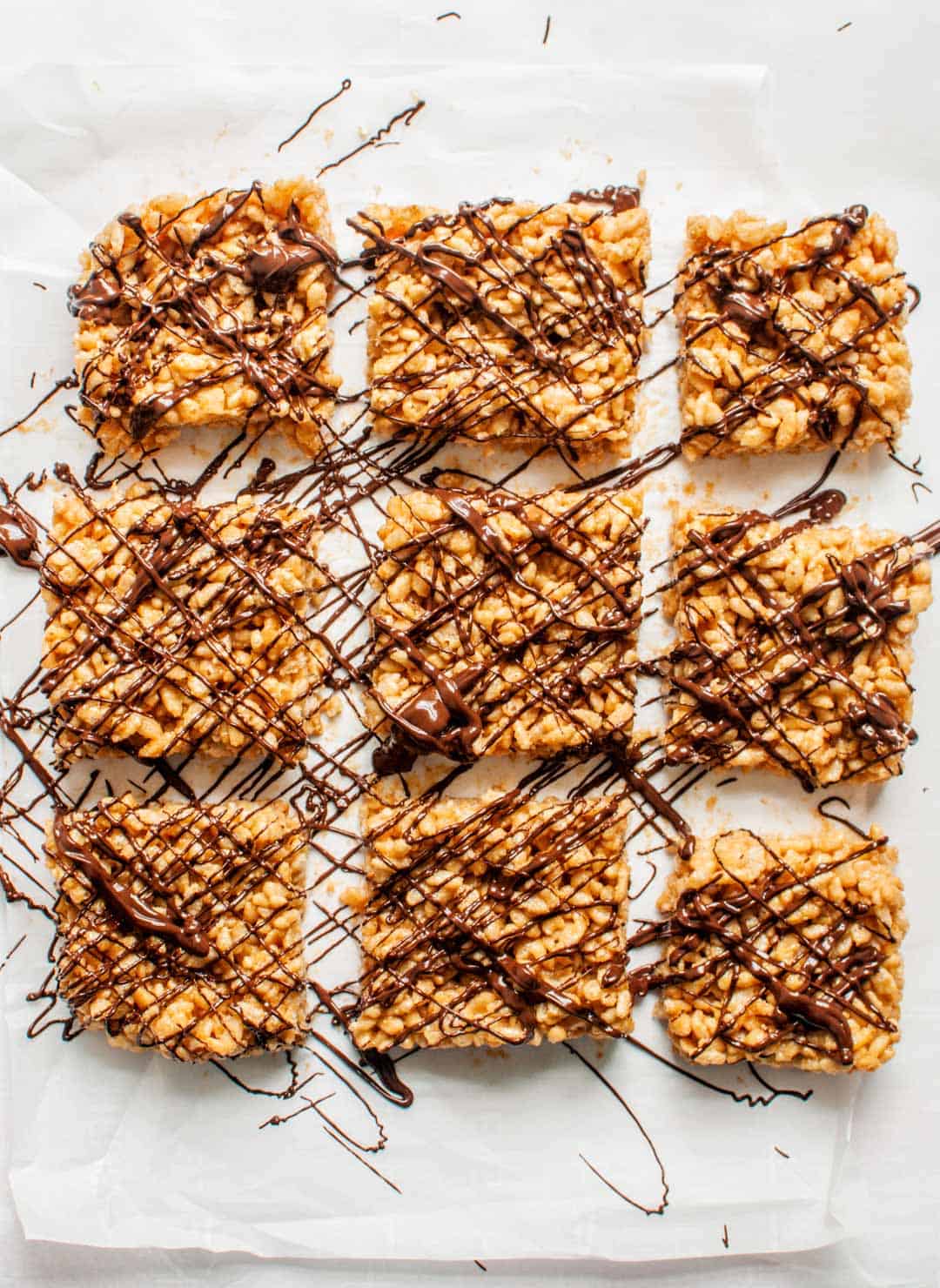 Peanut butter rice krispie treats with chocolate drizzle on parchment paper cut into nine pieces.