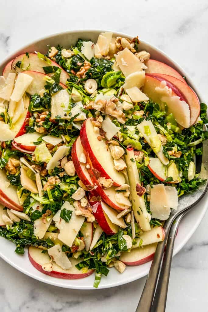An overhead shot of a white serving bowl with shredded brussels sprouts kale salad, topped with apples.
