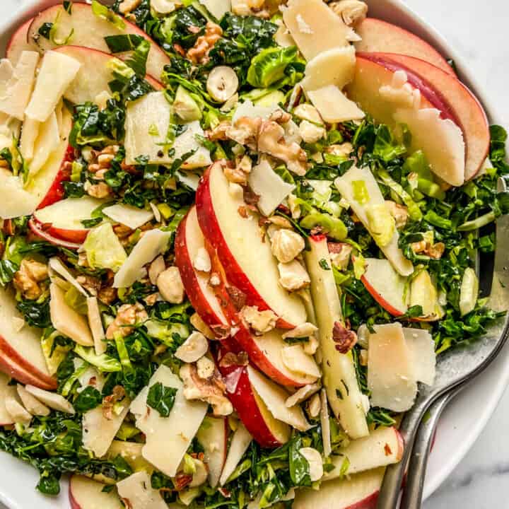 An overhead shot of a white serving bowl with shredded brussels sprouts kale salad, topped with apples.