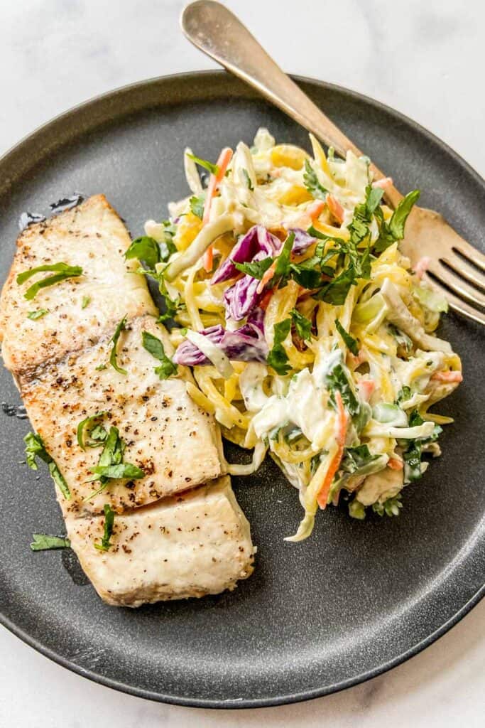 Closeup shot of a barramundi fillet next to mango slaw on a black plate with a fork.