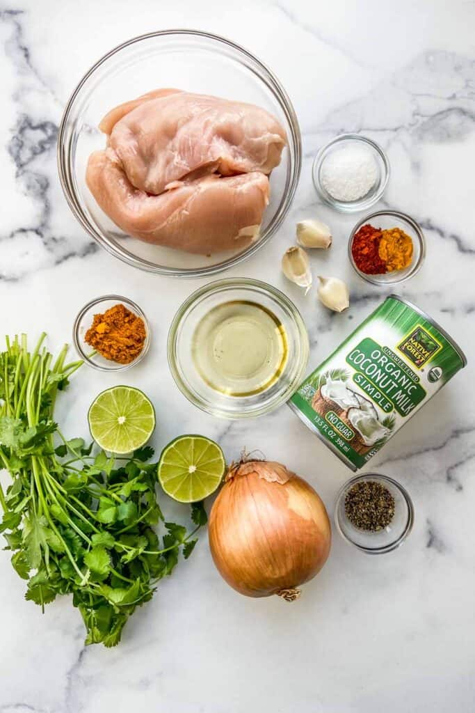 Ingredients for chicken curry on a marble background, including chicken coconut milk, a yellow onion, cilantro, lime, spices, garlic, and canola oil.