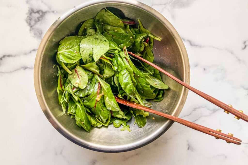 Green salad being tossed with dressing in a mixing bowl.