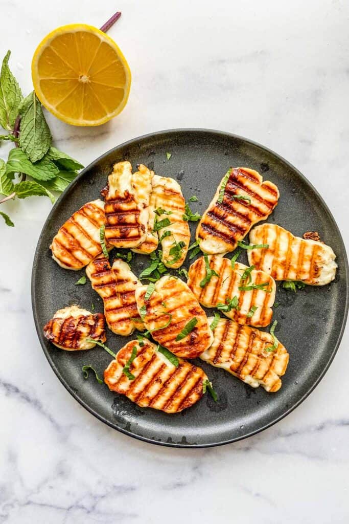 Slices of grilled halloumi cheese on a black plate with a lemon and pieces of mint to the side.