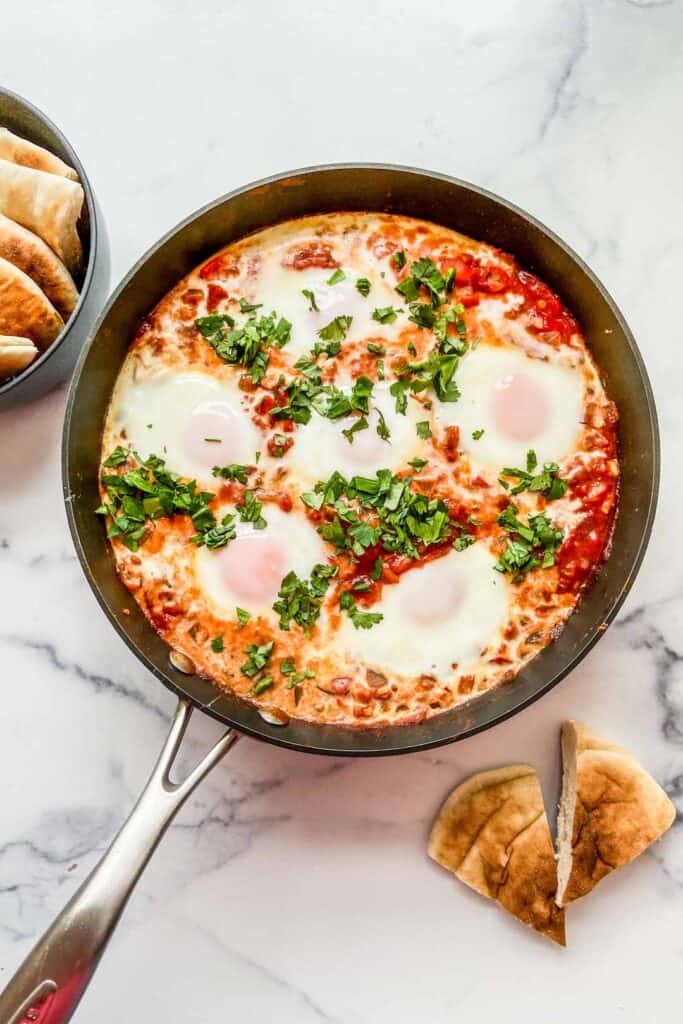 Overhead shot of shakshuka in a skillet with a bowl of cut up pita bread.