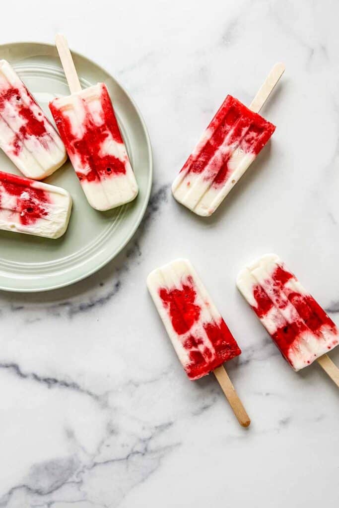 Six strawberry yogurt popsicles on a plate and marble slab.