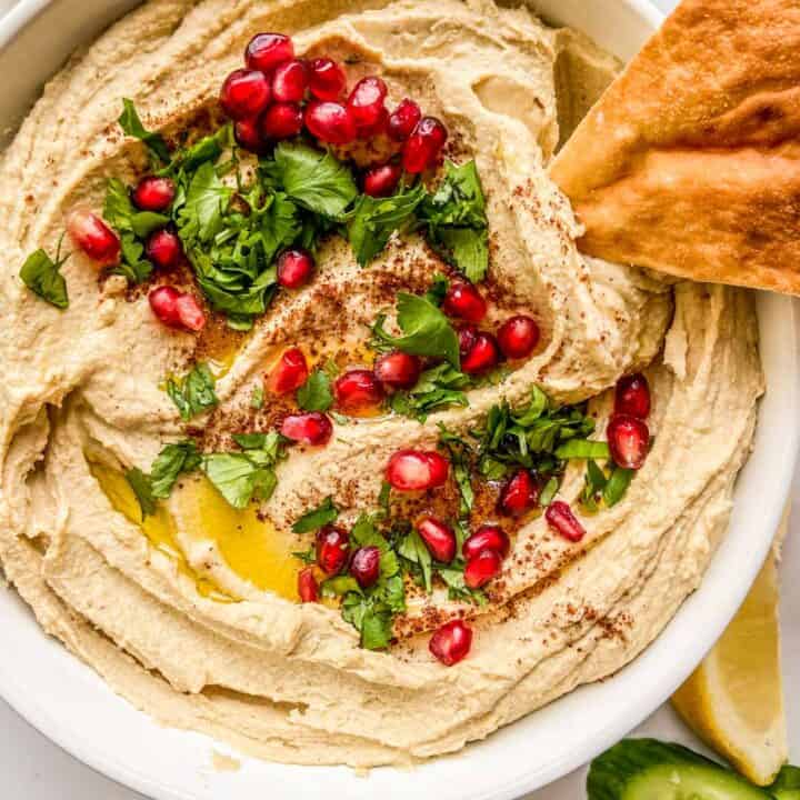A closeup shot of a bowl of hummus topped with olive oil, parsley, pomegranate seeds, and a piece of pita.