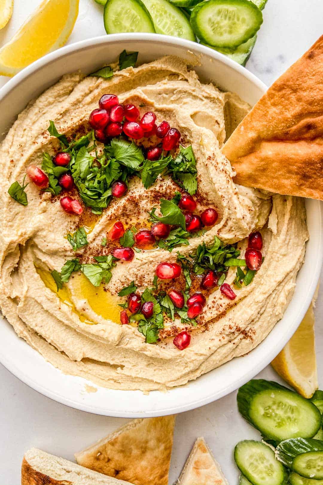 A closeup shot of a bowl of hummus topped with olive oil, parsley, pomegranate seeds, and a piece of pita.