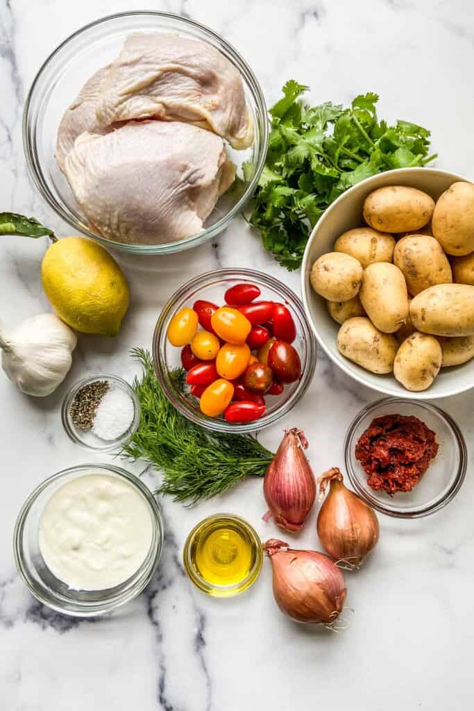 A bowl of chicken thighs, cherry tomatoes, small potatoes, dill, parsley, shallots, yogurt, lemon, garlic, olive oil, and harissa on a marble background.