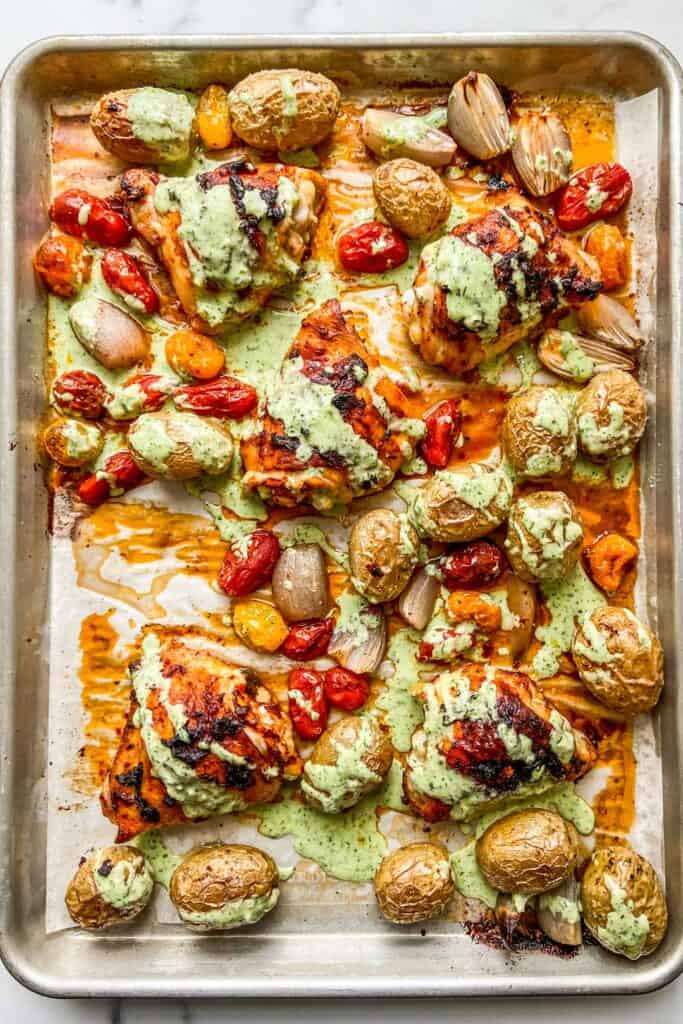 A sheet pan with roasted chicken, potatoes, tomatoes, and drizzled with a garlic yogurt sauce.