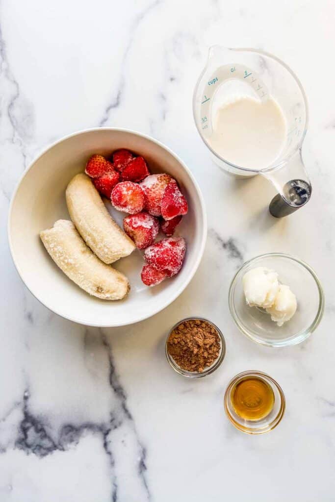 A bowl of frozen bananas and strawberries, a measuring cup with almond milk, a small bowl with coconut oil, a small bowl with cacao powder, and a small bowl with honey.