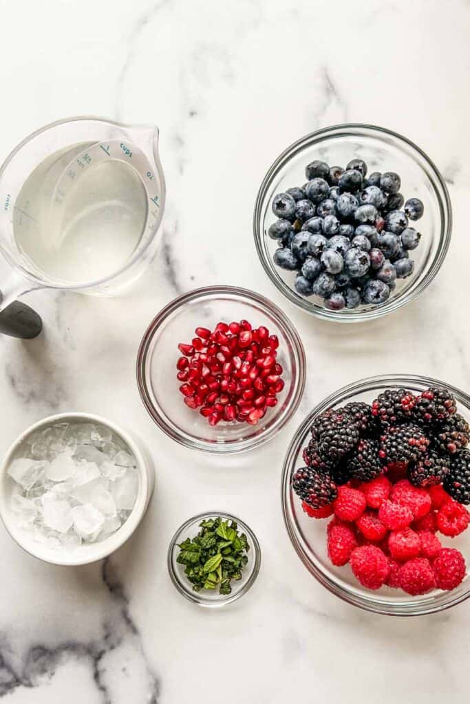 Ingredients for nature's cereal (blueberries, pomegranate seeds, raspberries, blackberries, mint, coconut water, and ice) on a marble background.