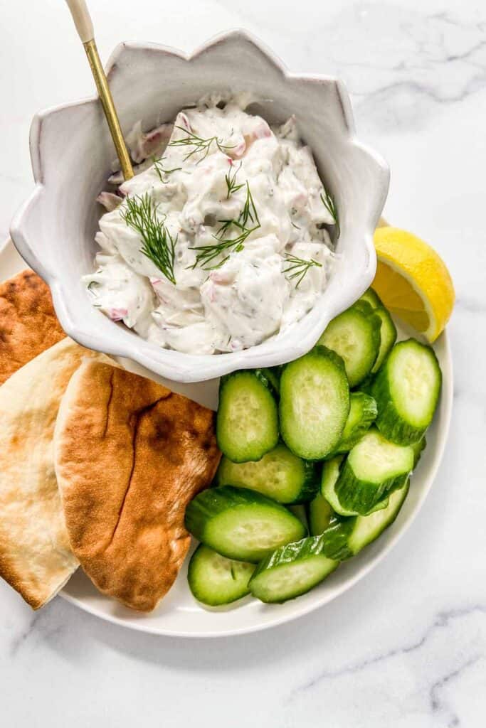 A bowl of radish tzatziki on a plate with chopped cucumbers and sliced pita bread.