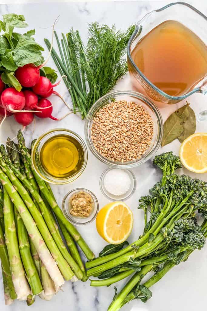 A grouping of ingredients, including radishes, asparagus, broccolini, vegetable stock, uncooked farro, olive oil, a halved lemon, bay leaves, salt, and mustard on a marble background.