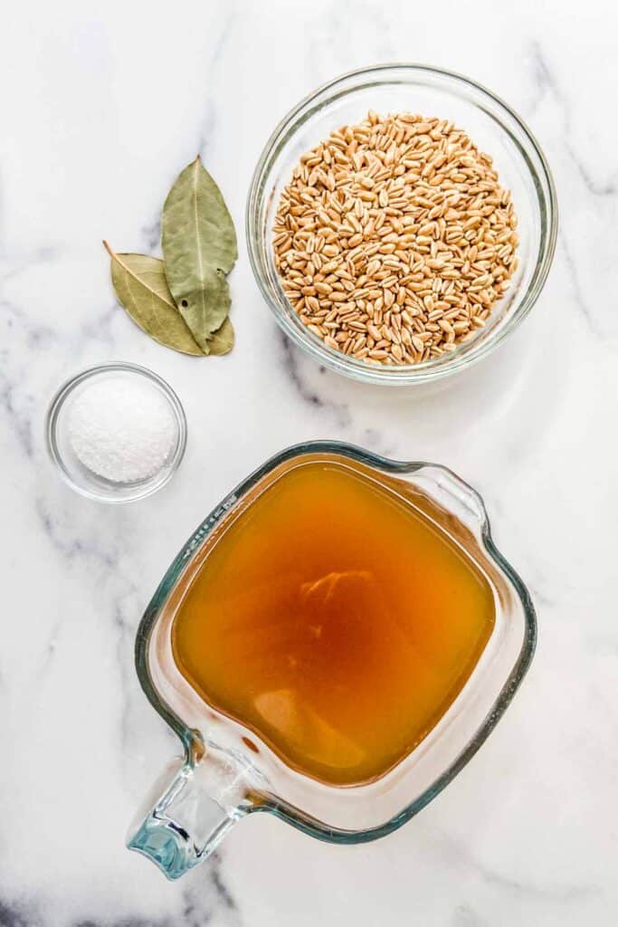 A measuring glass with vegetable stock, a bowl of farro, two bay leaves, and small bowl of kosher salt on a marble background.