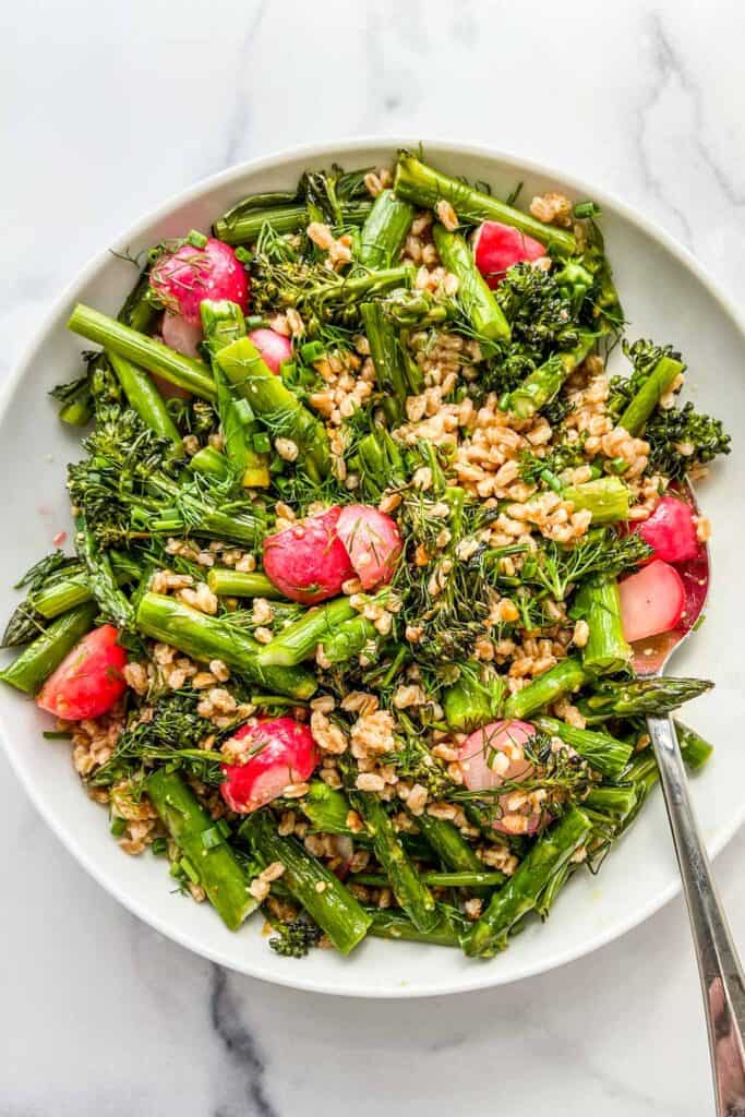 Roasted vegetables with farro in a large white serving bowl with a silver serving spoon.