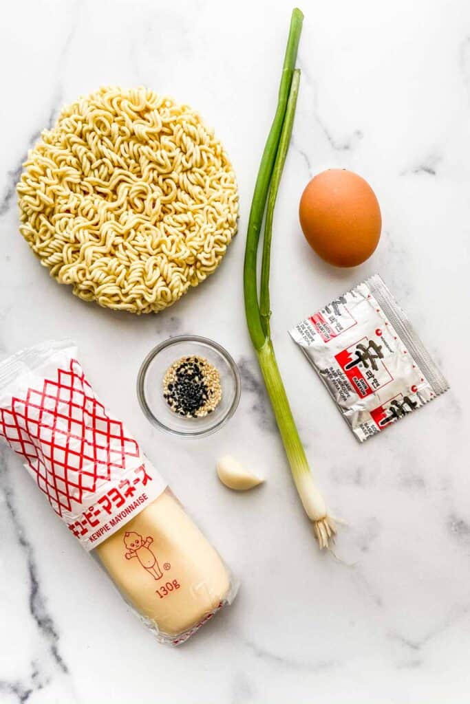 Ramen noodles, kewpie mayo, a green onion, an egg, a ramen spice packet, a clove of garlic, and a small bowl of sesame seeds on a marble background.