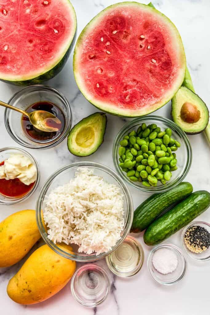 A halved watermelon, halved avocado, bowl of rice, bowl of edamame, green onion, cucumbers, mangoes, a bowl of marinating sauce, a bowl of sriracha mayo, and a bowl of sesame seeds on a marble background.