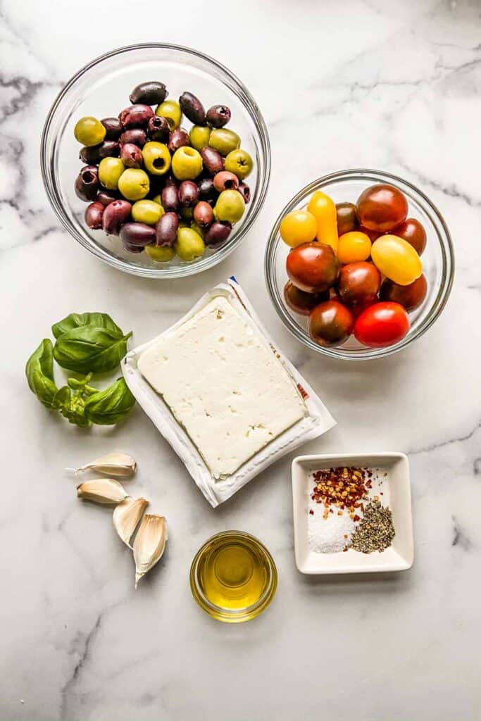 A bowl of olives, a bowl of cherry tomatoes, a block of feta, four garlic cloves, some basil leaves, a small bowl of olive oil, and some salt, pepper, and red pepper flakes. 