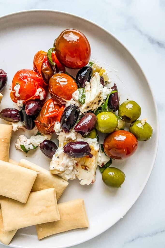 A plate with baked feta, olives, and tomatoes served alongside crackers.