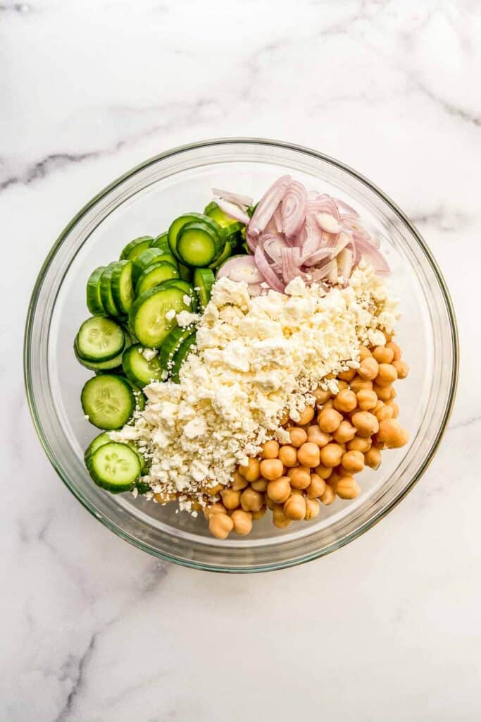 A large glass bowl with chickpeas, feta, cucumbers, and a chopped shallot.