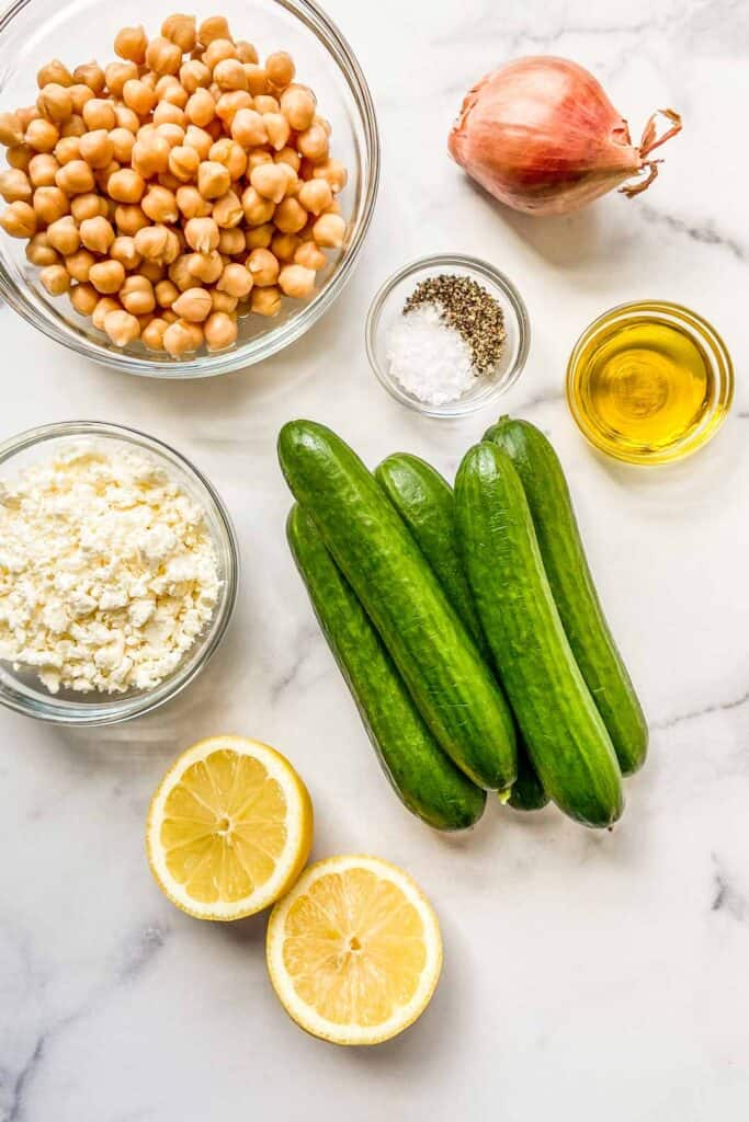 A bowl of chickpeas, several small cucumbers, a bowl of feta, a halved lemon, a bowl of olive oil, a shallot, and a bowl of salt and pepper.