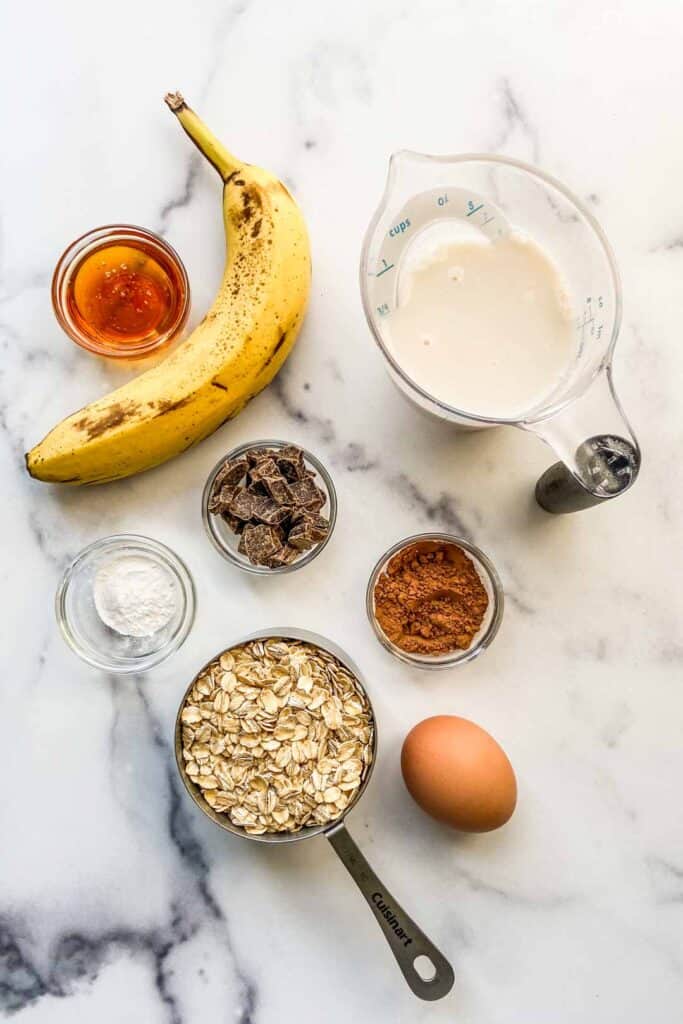 A banana, measuring cup of almond milk, bowl of honey, bowl of chocolate chunks, bowl of baking powder, bowl of cocoa powder, measuring cup of oats, and an egg sitting on a marble background.