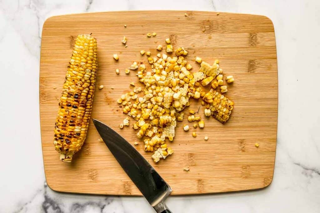 A cutting board with an ear of corn, a knife, and a pile of corn.