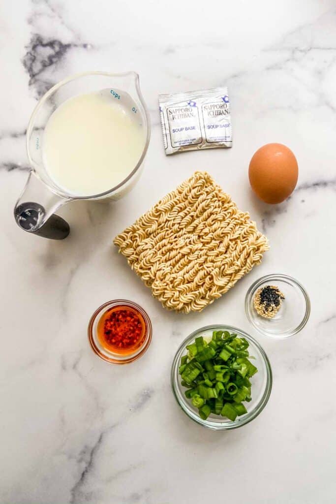 A measuring cup of milk, ramen noodles, an egg, chopped green onions, sesame seeds, and chili oil.