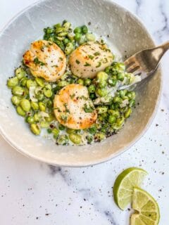 A bowl of edamame and pea sauce with three scallops on top and wedges of lime.