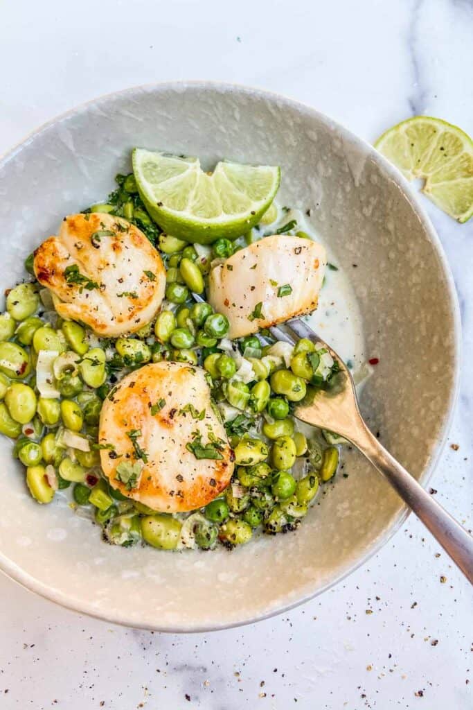 A bowl of edamame and pea sauce with three scallops on top and a wedge of lime.