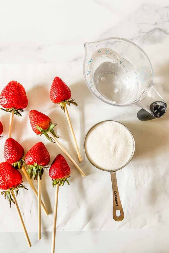 Strawberries on skewers, a cup of granulated sugar, and ½ cup of water in a plastic measuring cup.