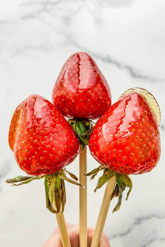 Three candied strawberries on wooden skewers.