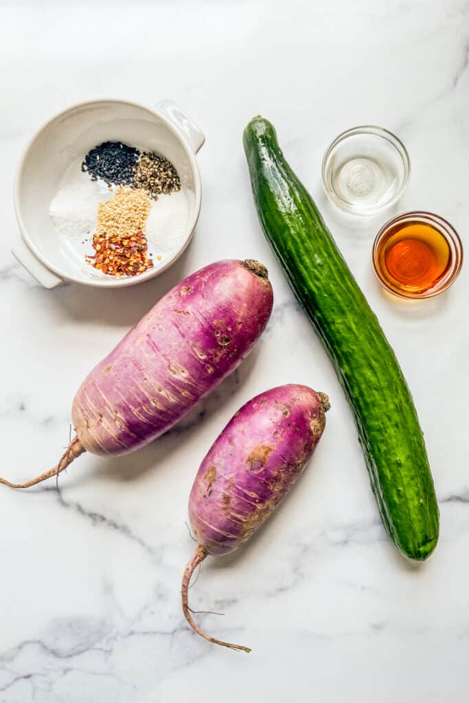 An English cucumber, two purple daikon radishes, a bowl of sesame oil, a bowl of rice wine vinegar, and a ramekin with sesame seeds and spices. 