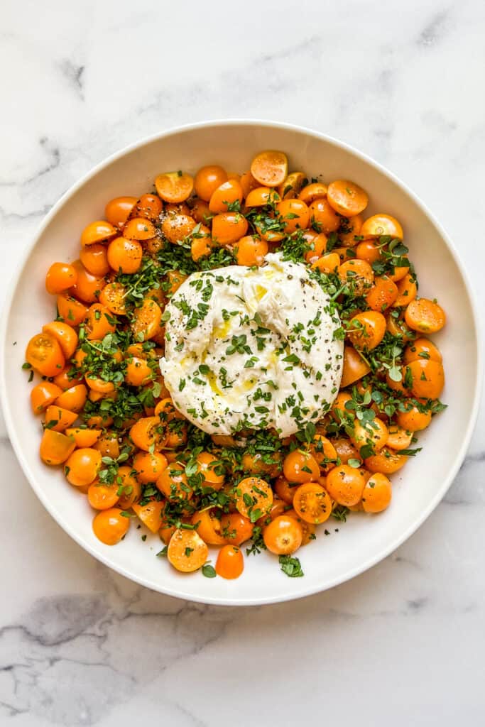 A large bowl with a ball of burrata in the middle surrounded by goldenberries and yellow cherry tomatoes, topped with chopped fresh herbs.