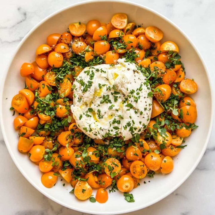 A large bowl with a ball of burrata in the middle surrounded by goldenberries and yellow cherry tomatoes, topped with chopped fresh herbs.