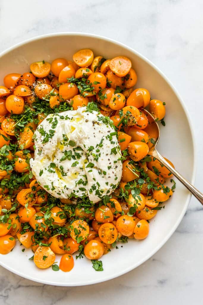 A large bowl with a ball of burrata in the middle surrounded by goldenberries and yellow cherry tomatoes, topped with chopped fresh herbs.