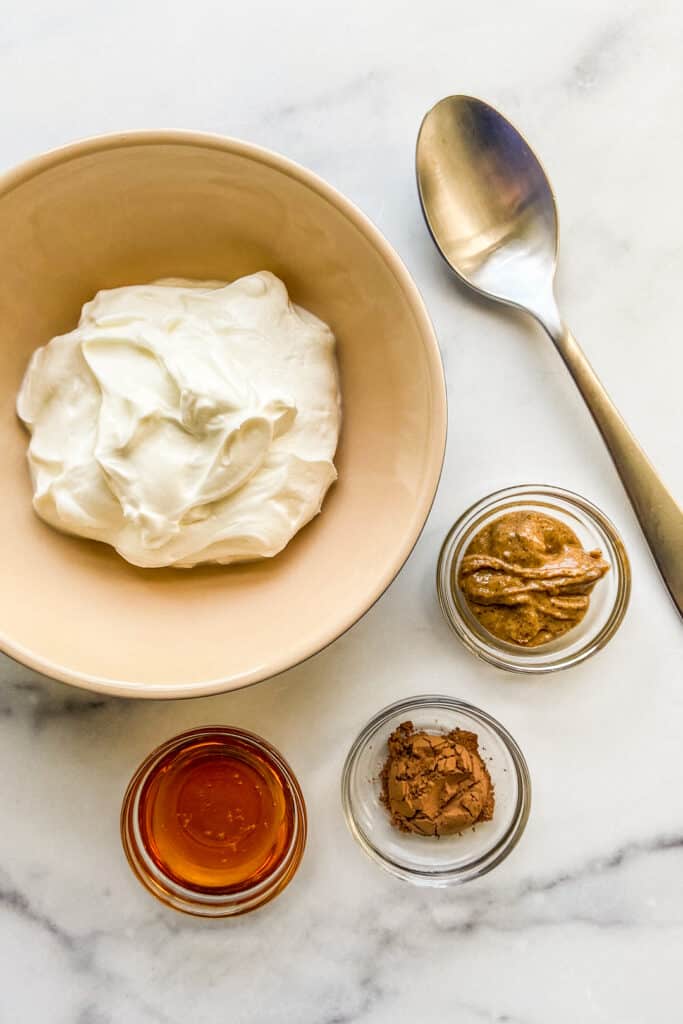 A bowl with Greek yogurt next to small bowls with honey, cocoa powder, and almond butter alongside a silver spoon.