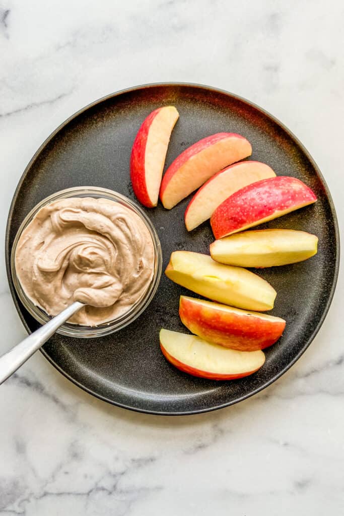 A small bowl of Greek yogurt dessert dip next to apple slices on a black plate.