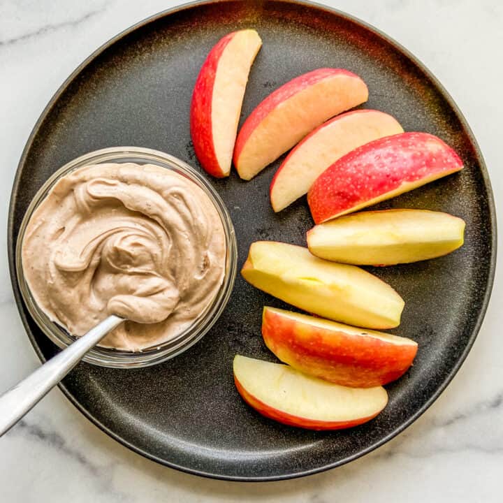 A small bowl of Greek yogurt dessert dip next to apple slices on a black plate.