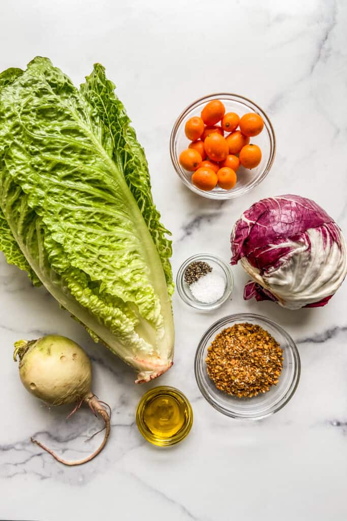 A head of lettuce, a bowl of kumquats, a head of raddichio, a bowl of dukkah, a bowl of olive oil, and a watermelon radish.