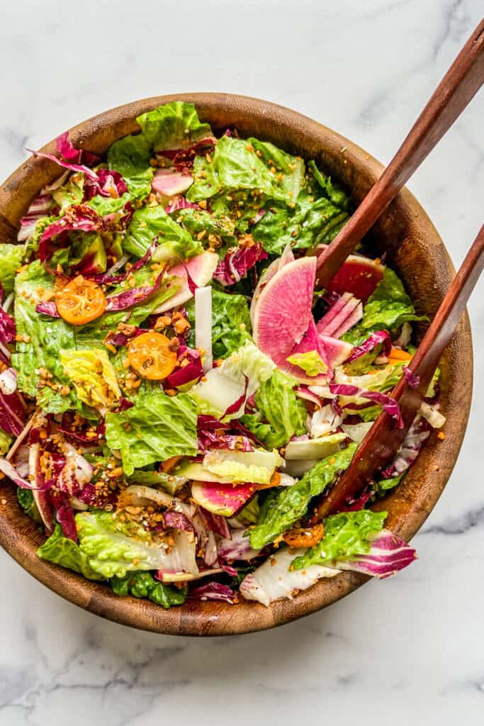 A large wooden salad bowl with a greens, radicchio, kumquats, watermelon radishes, and tongs. 